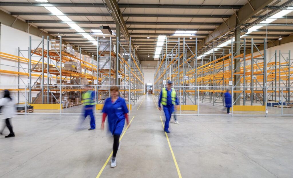 Trabalhadores caminhando em corredor de armazém industrial com estruturas de armazenamento, representando diferentes unidades operacionais
