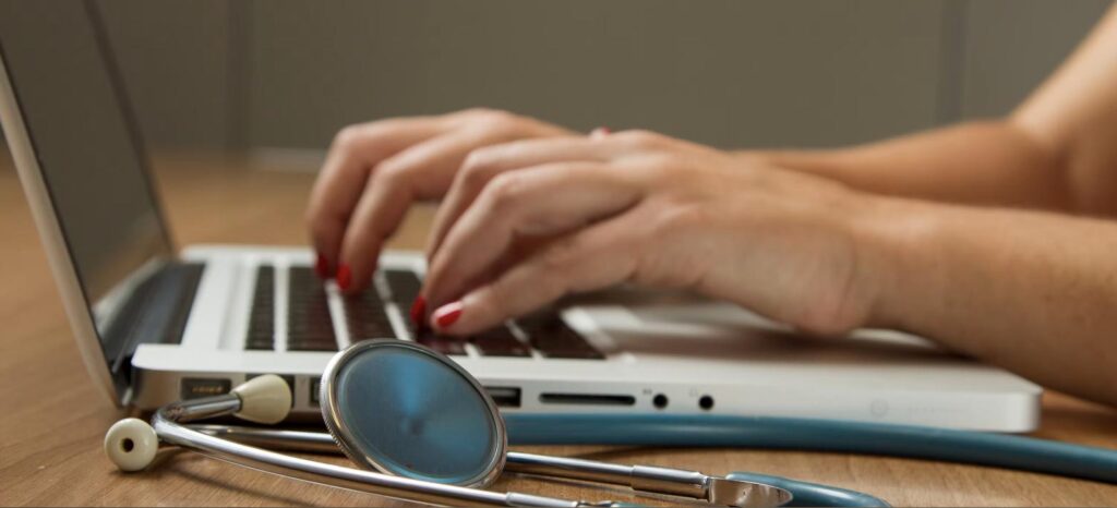 Mãos digitando em notebook ao lado de estetoscópio, simbolizando sigilo médico ocupacional